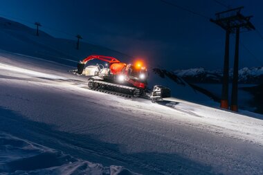 Eine beleuchtete Pistenraupe präpariert bei Nacht die Skipisten in Gargellen. Die Maschine fährt über die frisch verschneite Abfahrt, während im Hintergrund die Berggipfel und eine Seilbahnstütze zu sehen sind. Das Licht der Pistenraupe leuchtet den Schnee hell aus und schafft eine stimmungsvolle Atmosphäre. | © Montafon Tourismus GmbH, Marie Schilcher