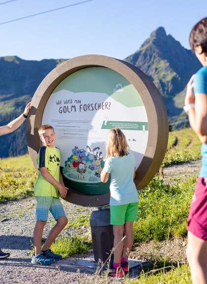 Eine funfköpfige Familie die am Erlebnisberg Golm auf Golmis Forschungspfad wandert und auf eine runde Tafel blickt auf der steht, wie man Golm Forscher wird | © Golm Silvretta Lünersee Tourismus GmbH Bregenz, Stefan Kothner