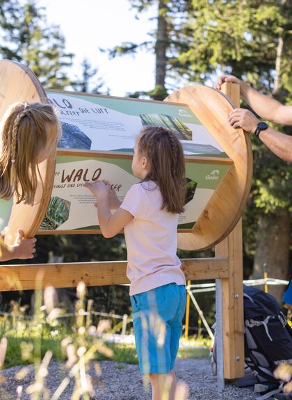 Ein Vater mit zwei Kindern auf Golmis Forschungspfad. Sie drehen gerade an einer rechteckigen Informationstafel mit vielen Informationen zum Wald. | © Golm Silvretta Lünersee Tourismus GmbH Bregenz, Stefan Kothner