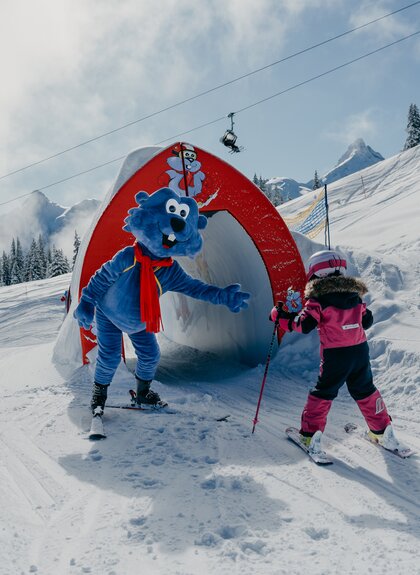 Maskottchen Golmi vom Erlebnisberg Golm mit einem Kind, dass am skifahren ist | © Golm Silvretta Lünersee Tourismus GmbH Bregenz, Philipp Schilcher