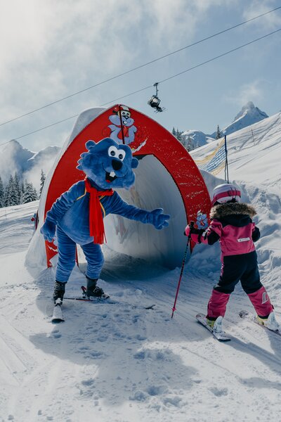 Maskottchen Golmi vom Erlebnisberg Golm mit einem Kind, dass am skifahren ist | © Golm Silvretta Lünersee Tourismus GmbH Bregenz, Philipp Schilcher